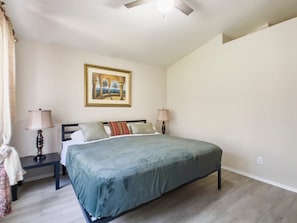 A bedroom with a bed, two bedside tables, a ceiling fan, a painting on the wall, and a window with curtains.
