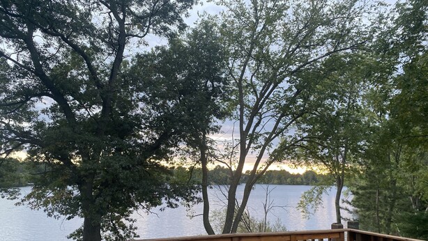 A wooden deck with a table and chairs, overlooking a calm lake with trees in the background.