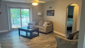 A spacious living room with a ceiling fan, a sofa, a coffee table, and a view through a sliding glass door to a balcony.