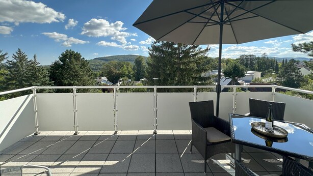Un balcón con mesa y sillas, un parasol grande, y vistas a un área boscosa y edificios a lo lejos.