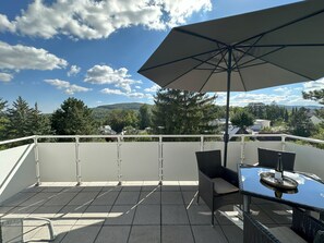 A balcony with a table and chairs, a large umbrella, and a view of a forested area and buildings in the distance.