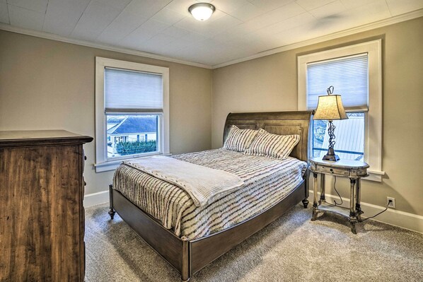 A bedroom with a wooden bed, a nightstand with a lamp, and two windows with blinds.