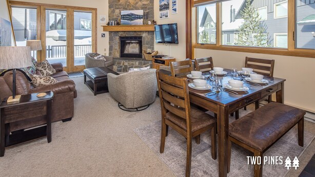 A spacious living room with a fireplace, a dining area set for four, and a large window offering a view of the outside. - Cozy Big Horn Home Walking Distance to Big Sky Resort | Ski-In/Out | Fireplace (Big Sky)