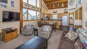 A cozy living room with a TV, comfortable seating, a kitchen area, and large windows offering views of the outdoors. - Cozy Big Horn Home Walking Distance to Big Sky Resort | Ski-In/Out | Fireplace (Big Sky)