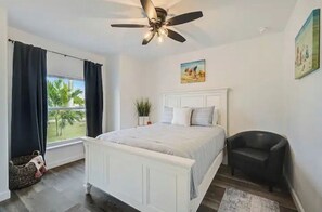 A bedroom with a bed, a chair, a ceiling fan, a window with curtains, and a painting on the wall.