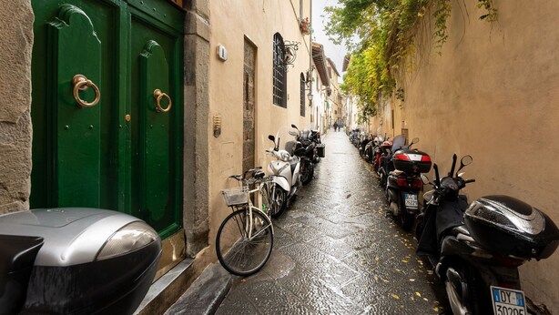 Una calle estrecha con scooters y bicicletas aparcados, puertas verdes y un edificio con techo de tejas.