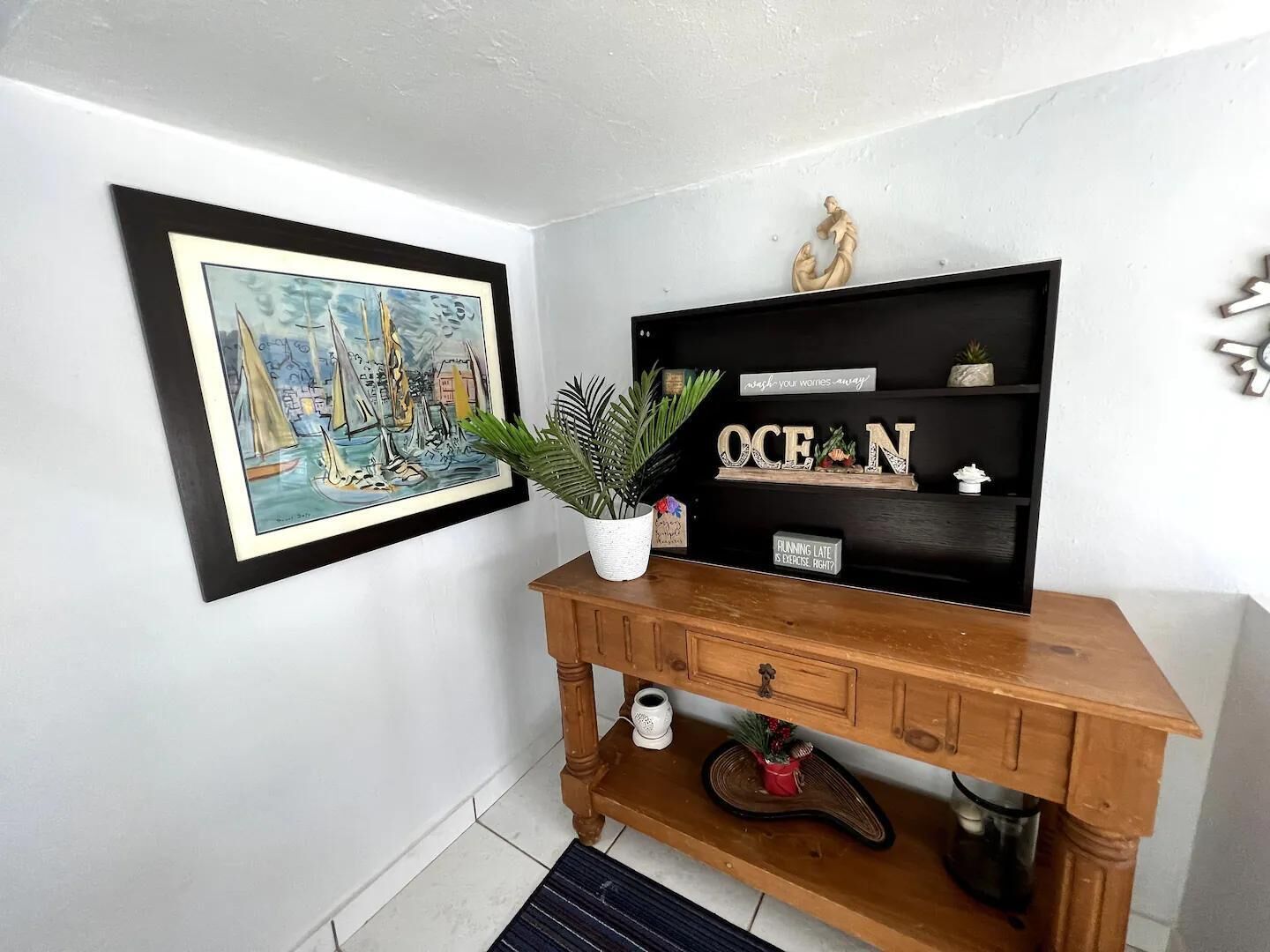A wooden console table with a potted plant, a framed painting of sailboats, and decorative items including a sign that says "OCEAN" and another that says "FLANNING LATE IS EXERCISE, RIGHT?".