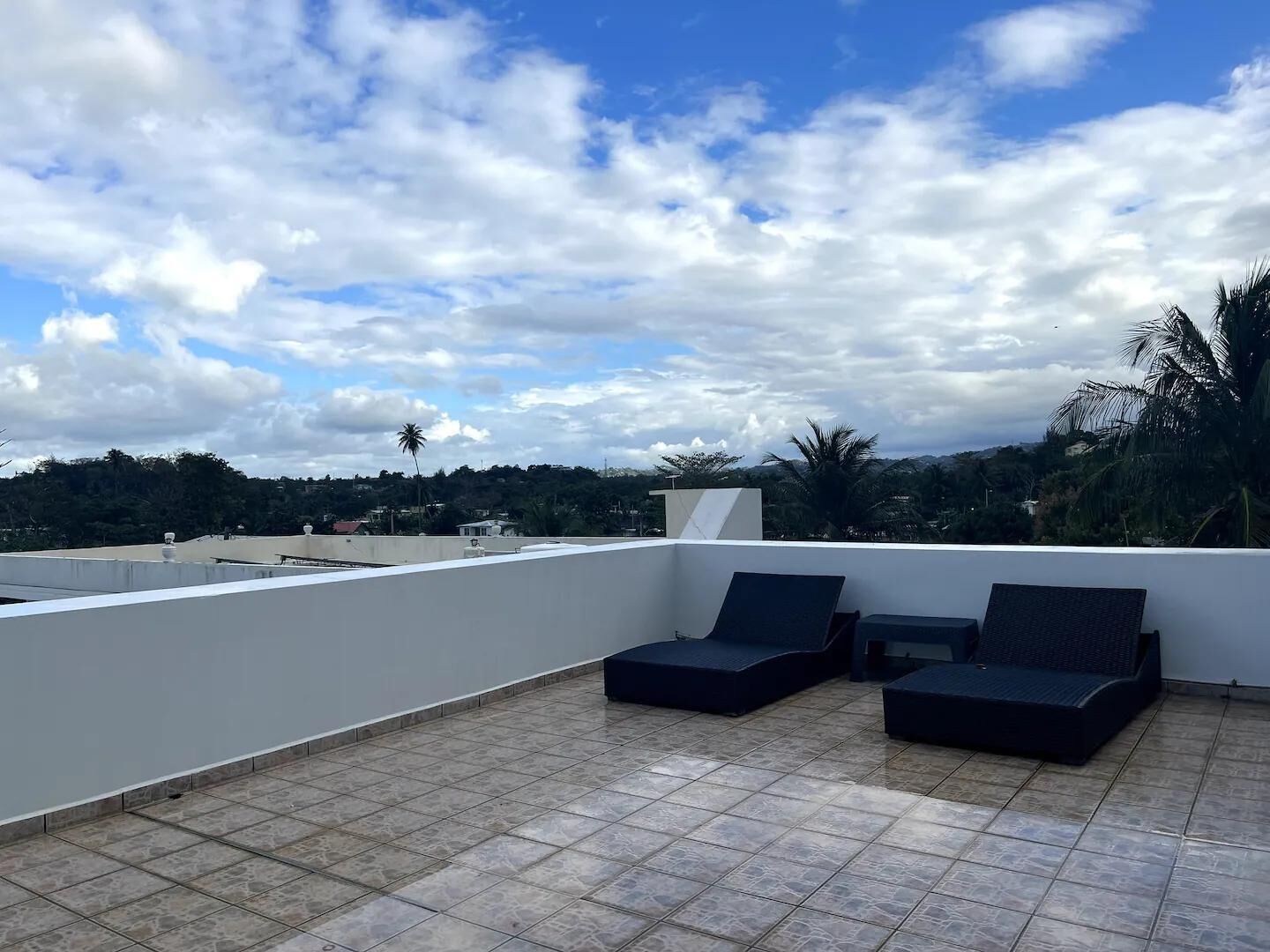 Una terraza en la azotea con dos sillones oscuros y una pequeña mesa, rodeada por un muro blanco y un suelo de baldosas, con vistas a un paisaje de árboles y edificios bajo un cielo parcialmente nublado.