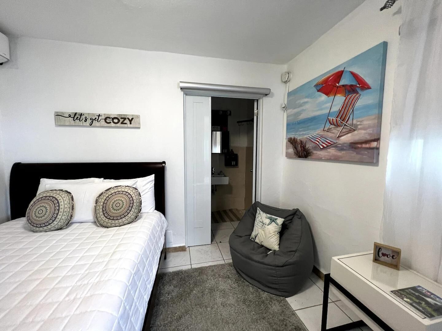 A bedroom with a bed, a bean bag chair, a painting of a beach scene, and a sign that says "let's get cozy".