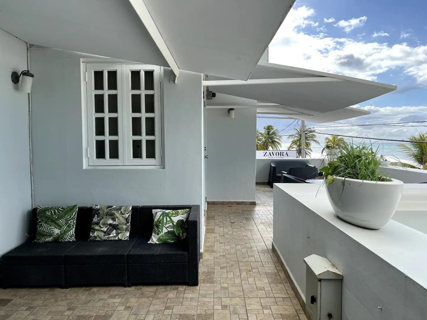 A balcony with a white wall, a black sofa with patterned cushions, a potted plant, and a view of the ocean.
