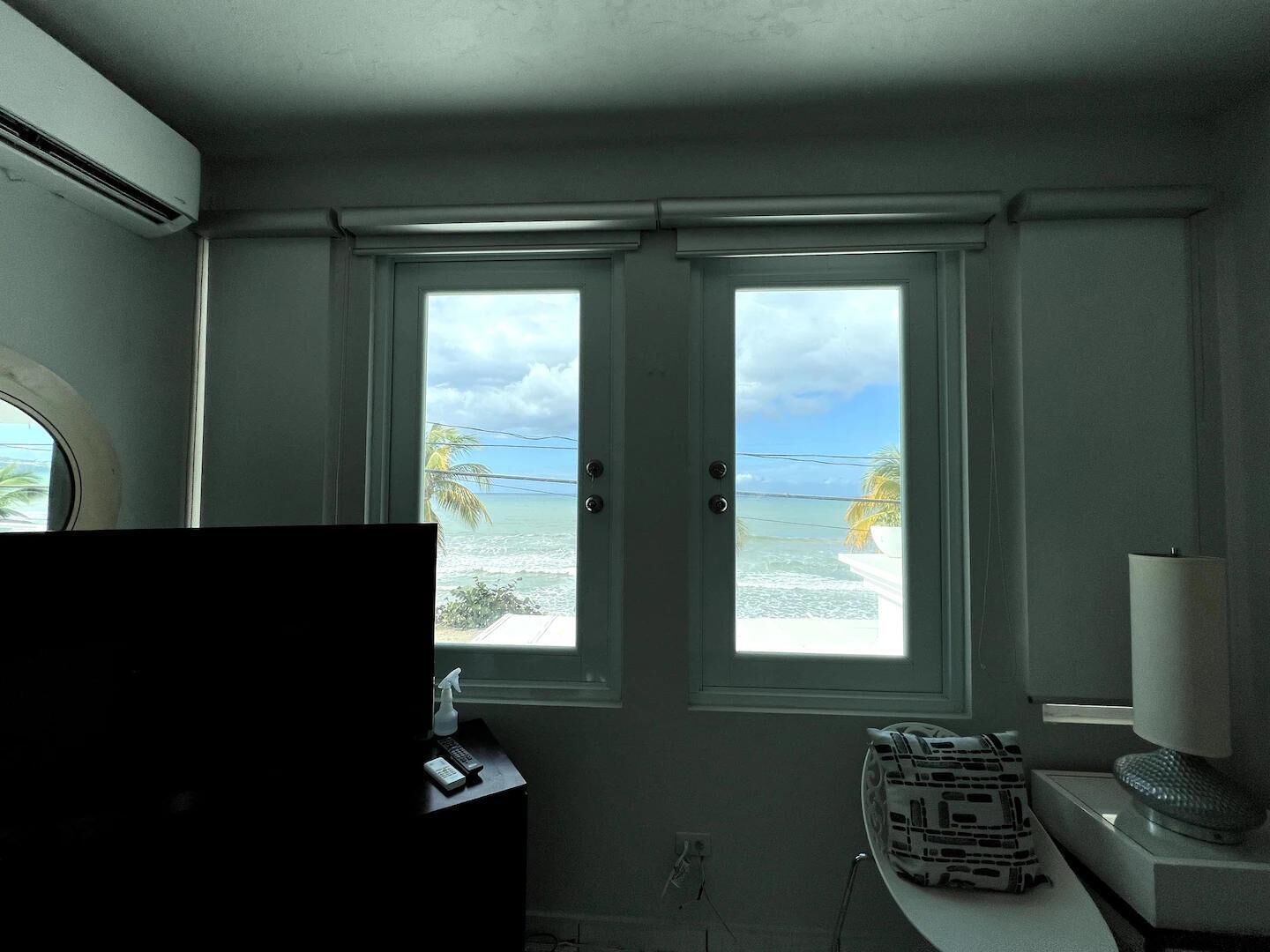 Et værelse med et stort vindue der udover en strand, et fjernsyn, og et lille bord med en lampe og en pynteting.