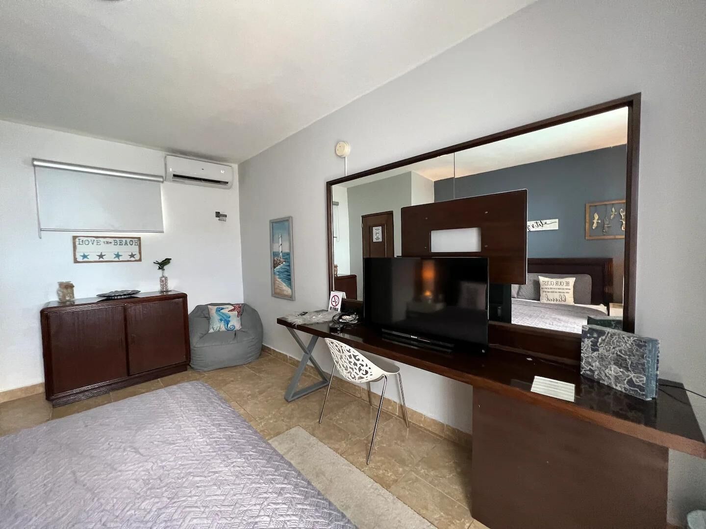 A hotel room with a bed, a desk with a flat-screen TV, a chair, a dresser, and a large mirror.