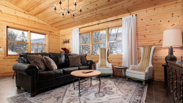 Un interior acogedor de cabaña de madera con un sofá de cuero, una mesita redonda y un sillón blanco.