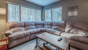 A corner living room with a large L-shaped sectional sofa, a wooden coffee table, and a large window with a view of trees.