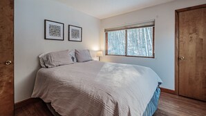 A bedroom with a large bed, two framed artworks on the wall, a window with a view of trees, and a wooden wardrobe.