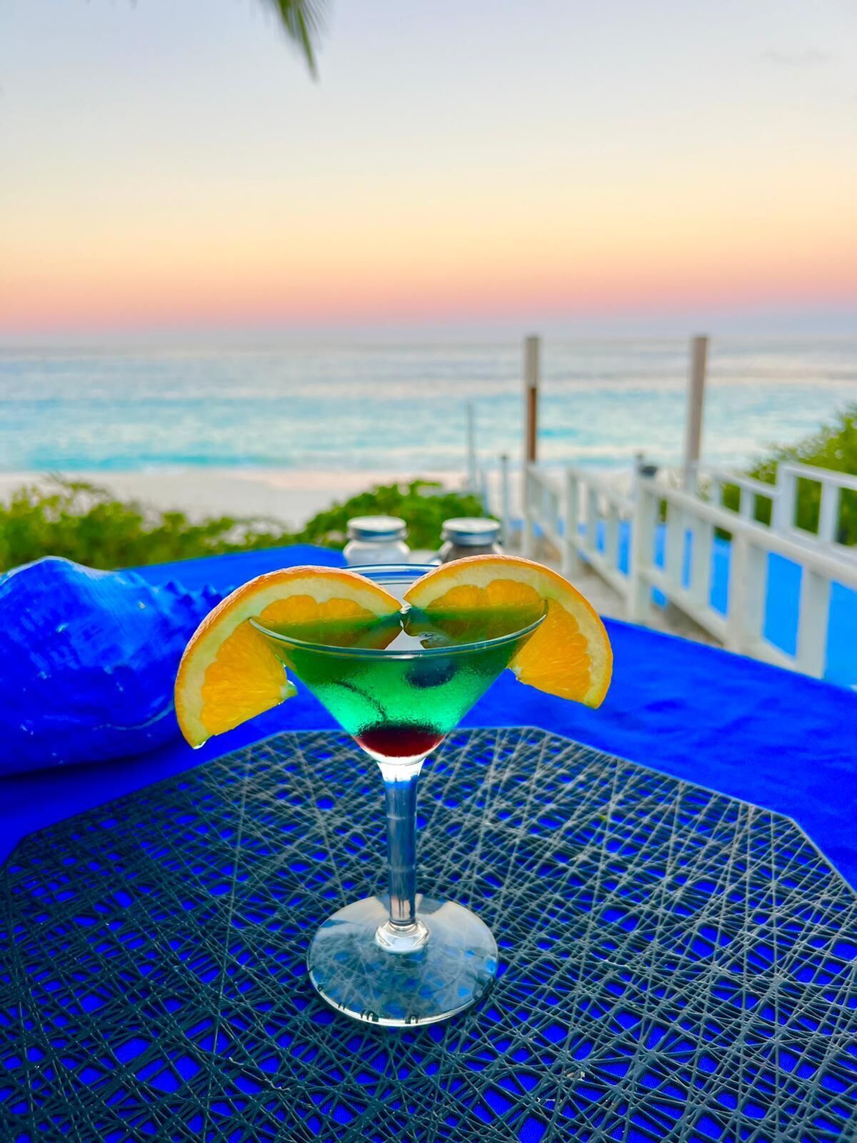 Ein Cocktail auf einem blauen Tisch mit Blick auf den Sonnenuntergang über dem Meer.