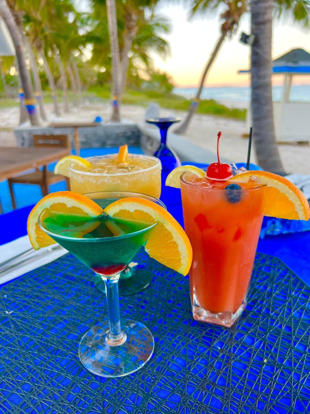 Three cocktails on a blue table with a beach view in the background.