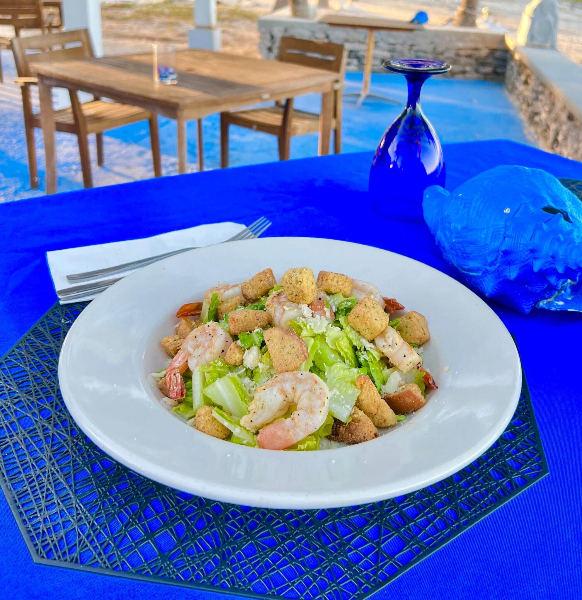 A seafood salad on a white plate with croutons and lettuce, set on a blue tablecloth with a blue umbrella in the background.