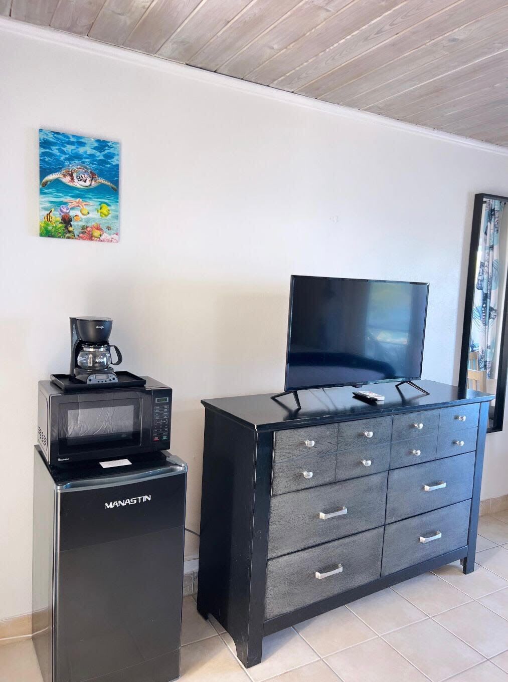 A black microwave and coffee maker on a small cabinet, a black TV on a dresser, and a painting of an underwater scene on the wall.