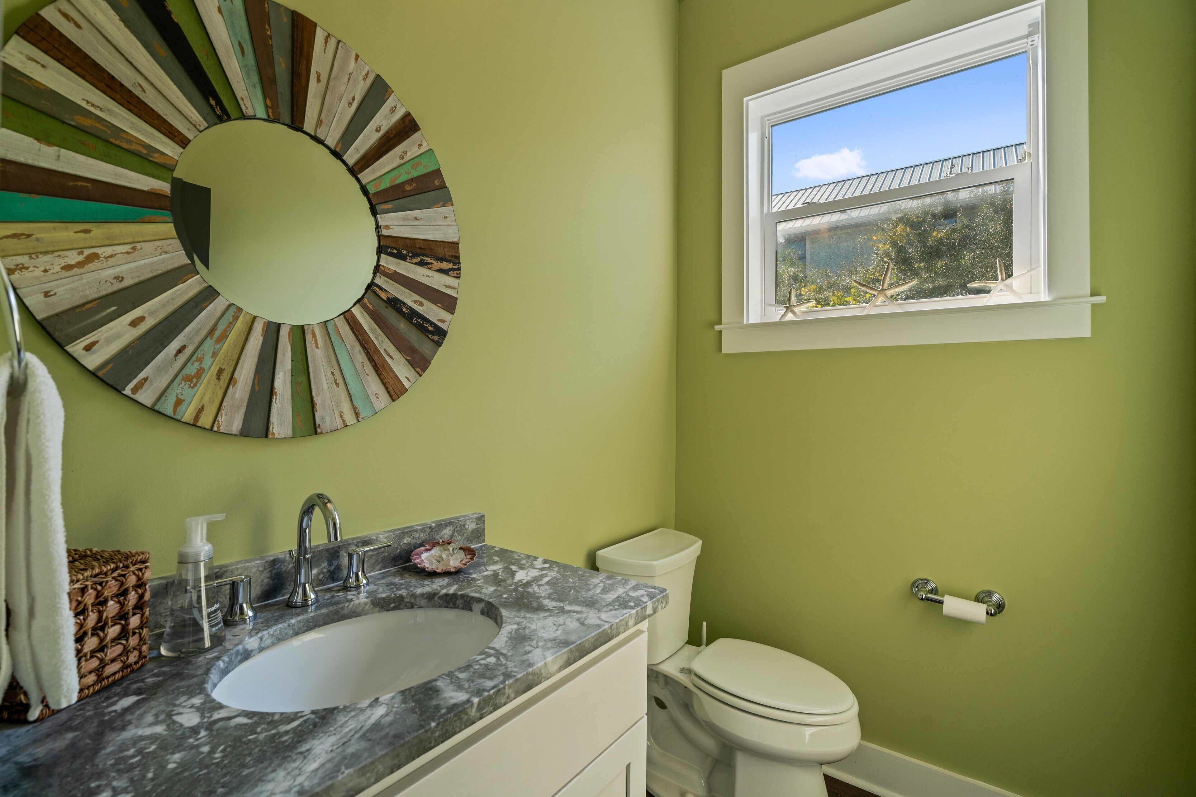 A bathroom with a marble sink, a toilet, a window with a view of the outside, and a circular mirror on the wall.