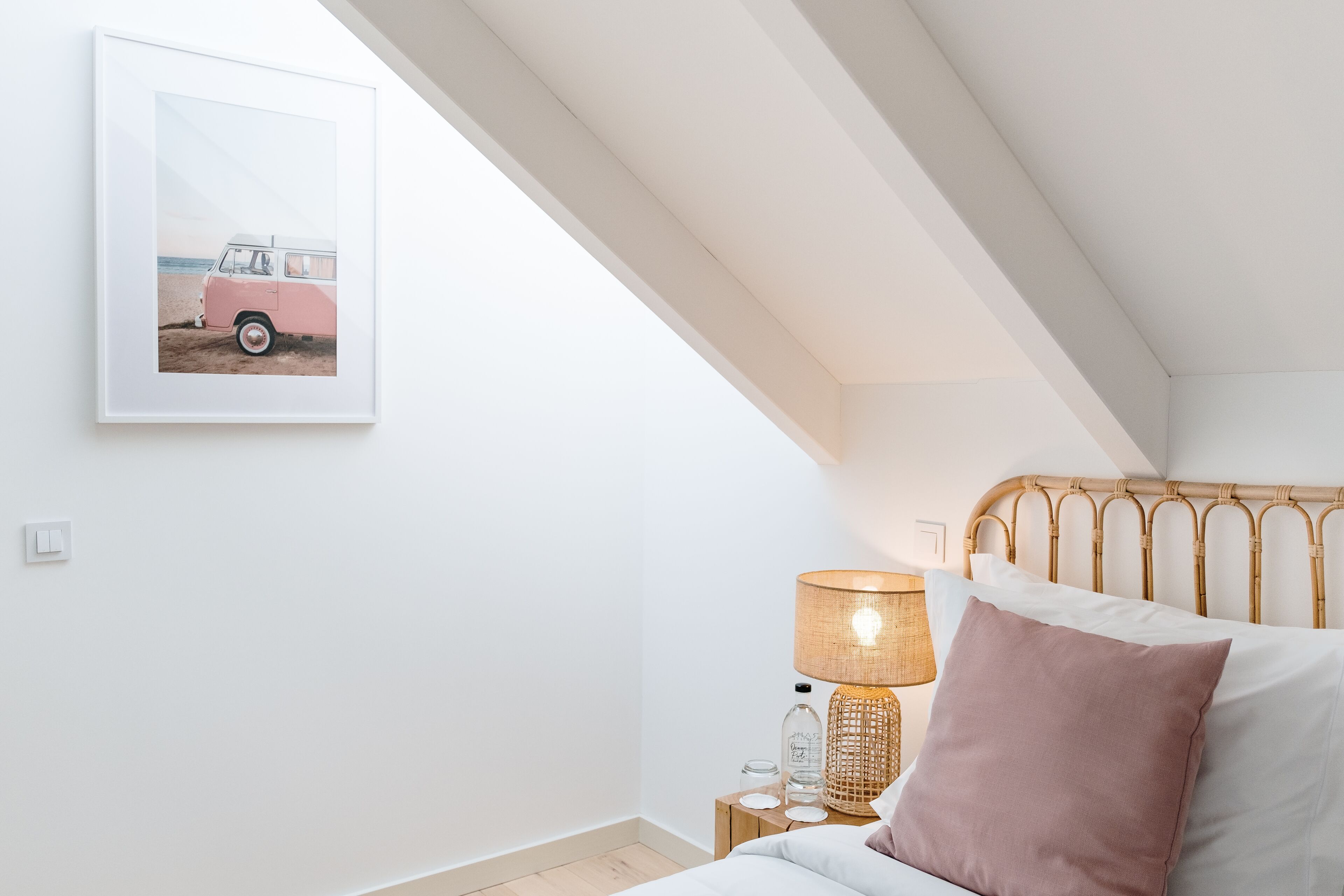 Ein Schlafzimmer mit weißer Wand, einem gerahmten Bild eines pinkfarbenen Lieferwagens, einem Nachttisch mit Lampe und einem Bett mit Holz-Kopfstück.