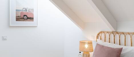 A bedroom with a white wall, a framed picture of a pink van, a bedside table with a lamp, and a bed with a wooden headboard.