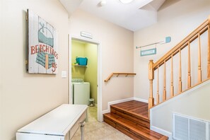 A staircase with wooden handrails and a 'Welcome' sign, leading to a laundry area with a washing machine and a shelf with a blue bag.