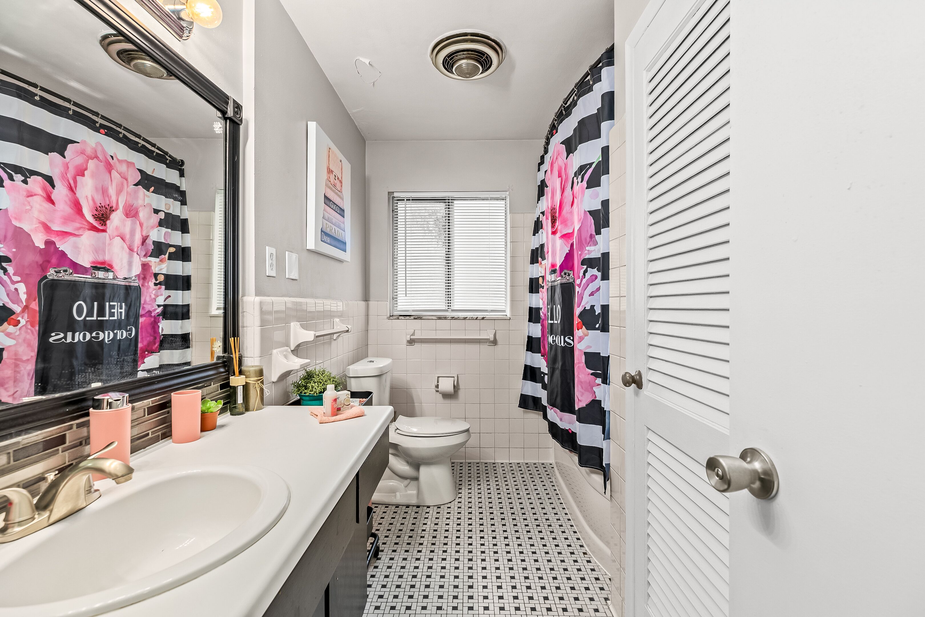 A bathroom with a white sink, toilet, and window. There are pink and black floral curtains, and a black and white patterned shower curtain.
