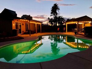 A swimming pool with surrounding patio seating and a covered area, surrounded by palm trees and a sunset sky. - Very own Arizona retreat with private pool and putting green. Sleeps six (Chandler)