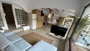 A modern living room with a sofa, a television, and a kitchen area with a refrigerator and sink. - Costa Maresme 20 km BCN, Garden apartment & Private pool (Cabrils)
