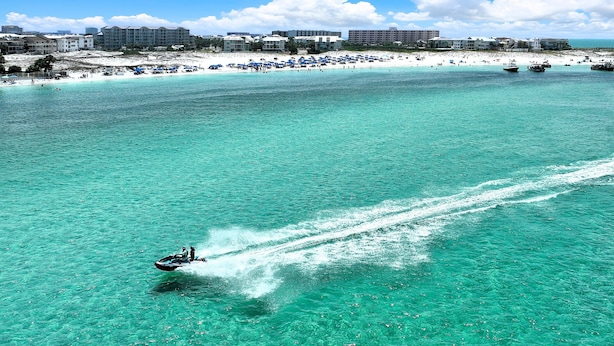 Una playa con agua turquesa cristalina, un jet ski y una hilera de edificios al fondo.