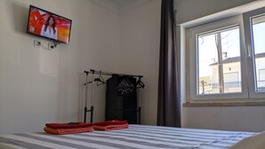 A hotel room with a bed, a TV mounted on the wall, a window with a view of buildings, and a drying rack with clothes. - GuestHouse Marialva Park (Seixal)