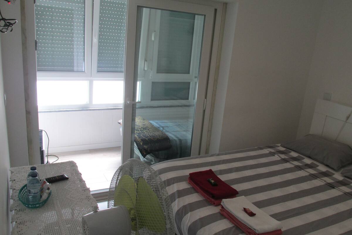Hotelkamer met een bed, een tafel met een fles en een afstandsbediening, een raam met zonwering, en uitzicht op een balkon.