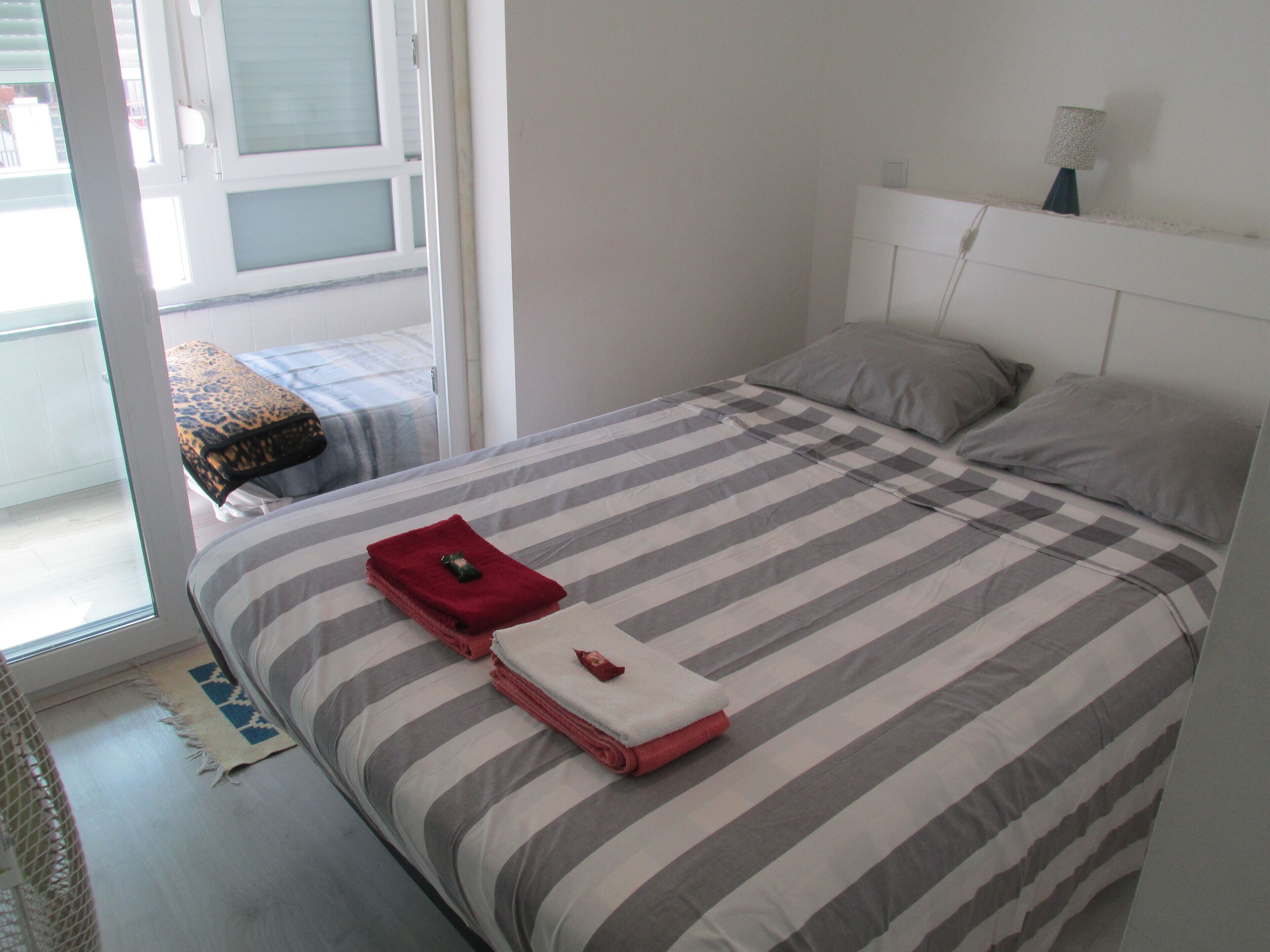 Uma cama bem arrumada com roupa de cama listrada, uma pequena mesa de cabeceira e uma janela com persianas.