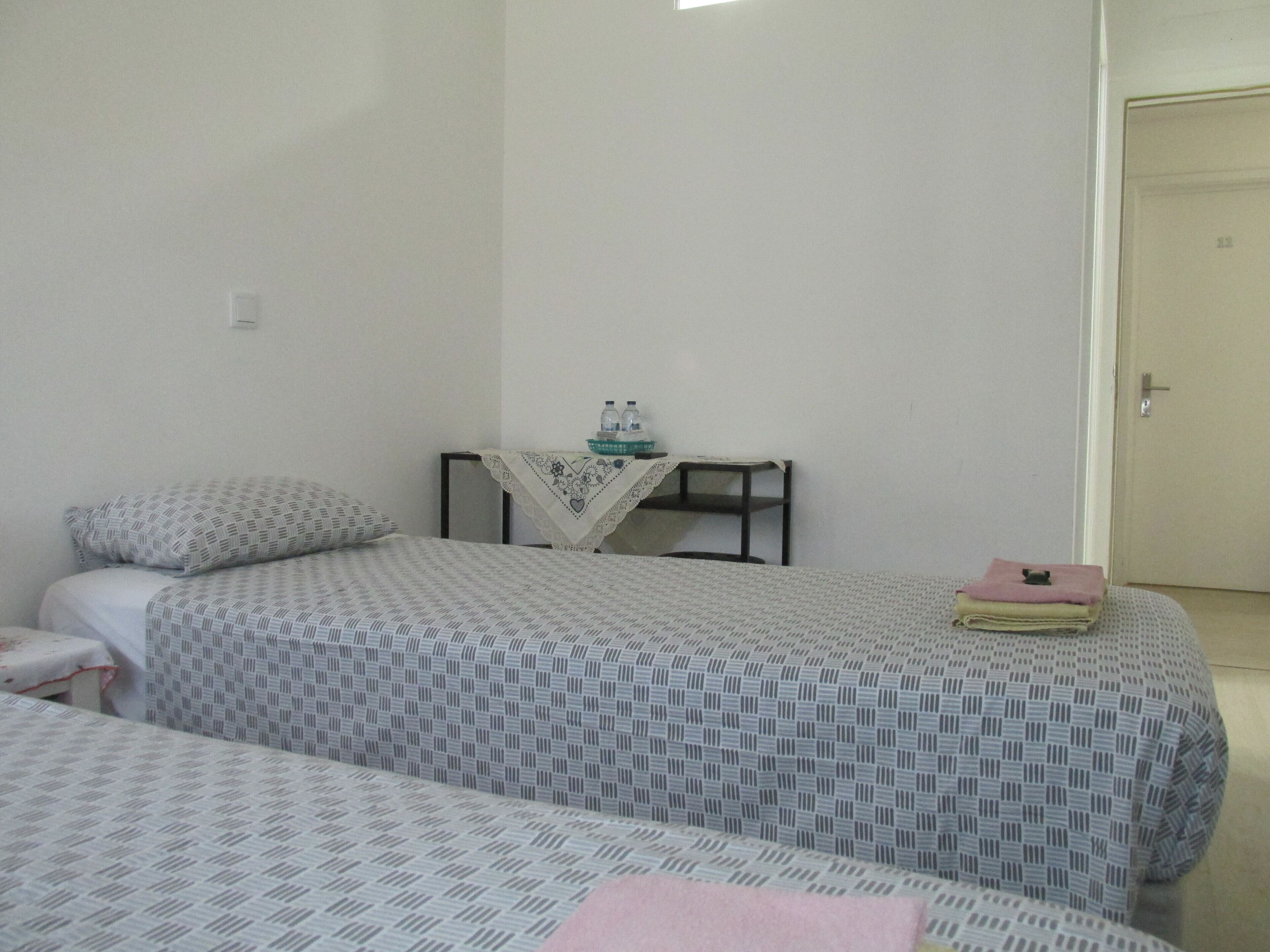 A simple room with two beds, a small table with a lace tablecloth, and a door.