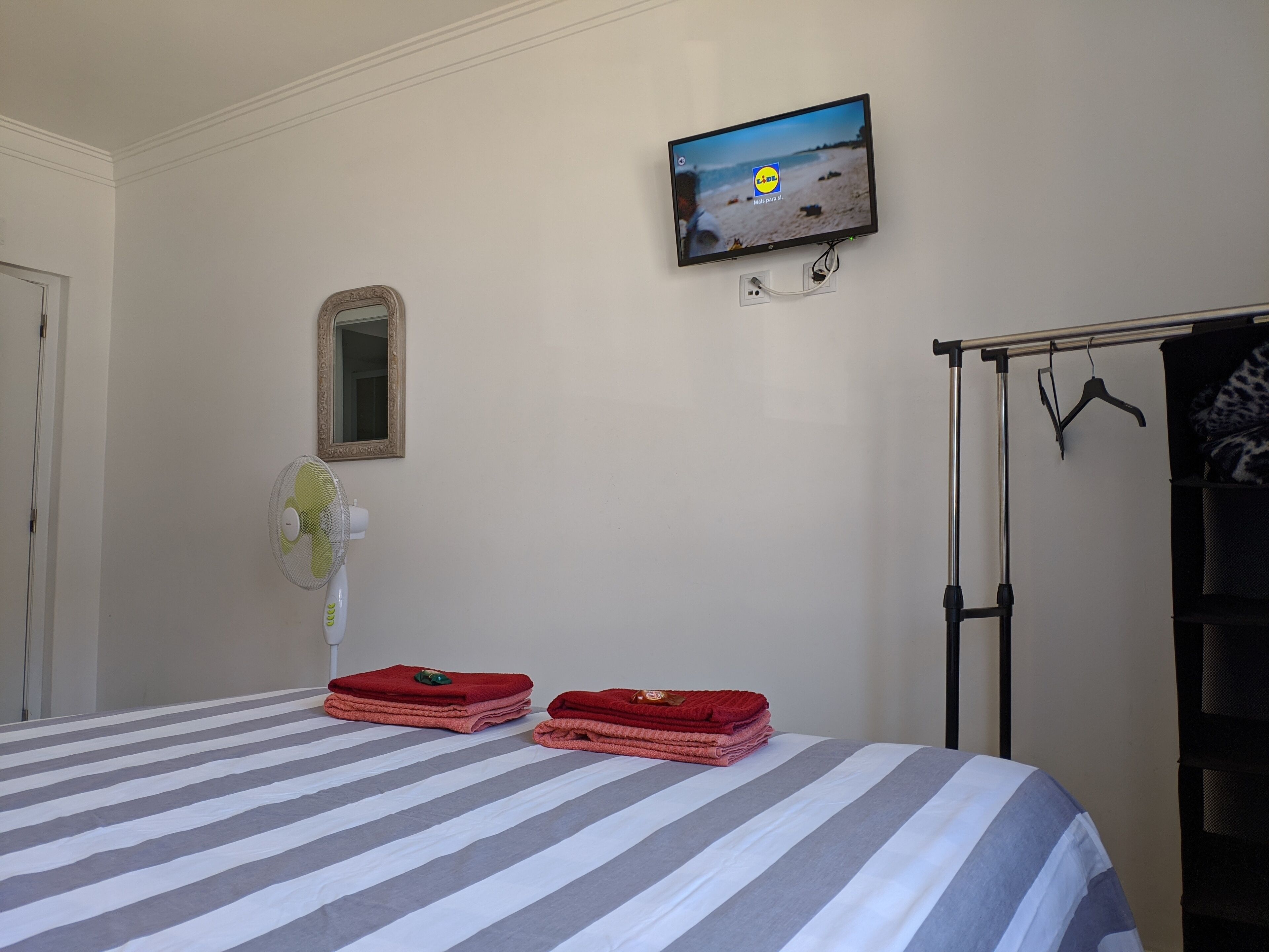 Una habitación de hotel con cama, televisor montado en la pared, espejo y perchero.