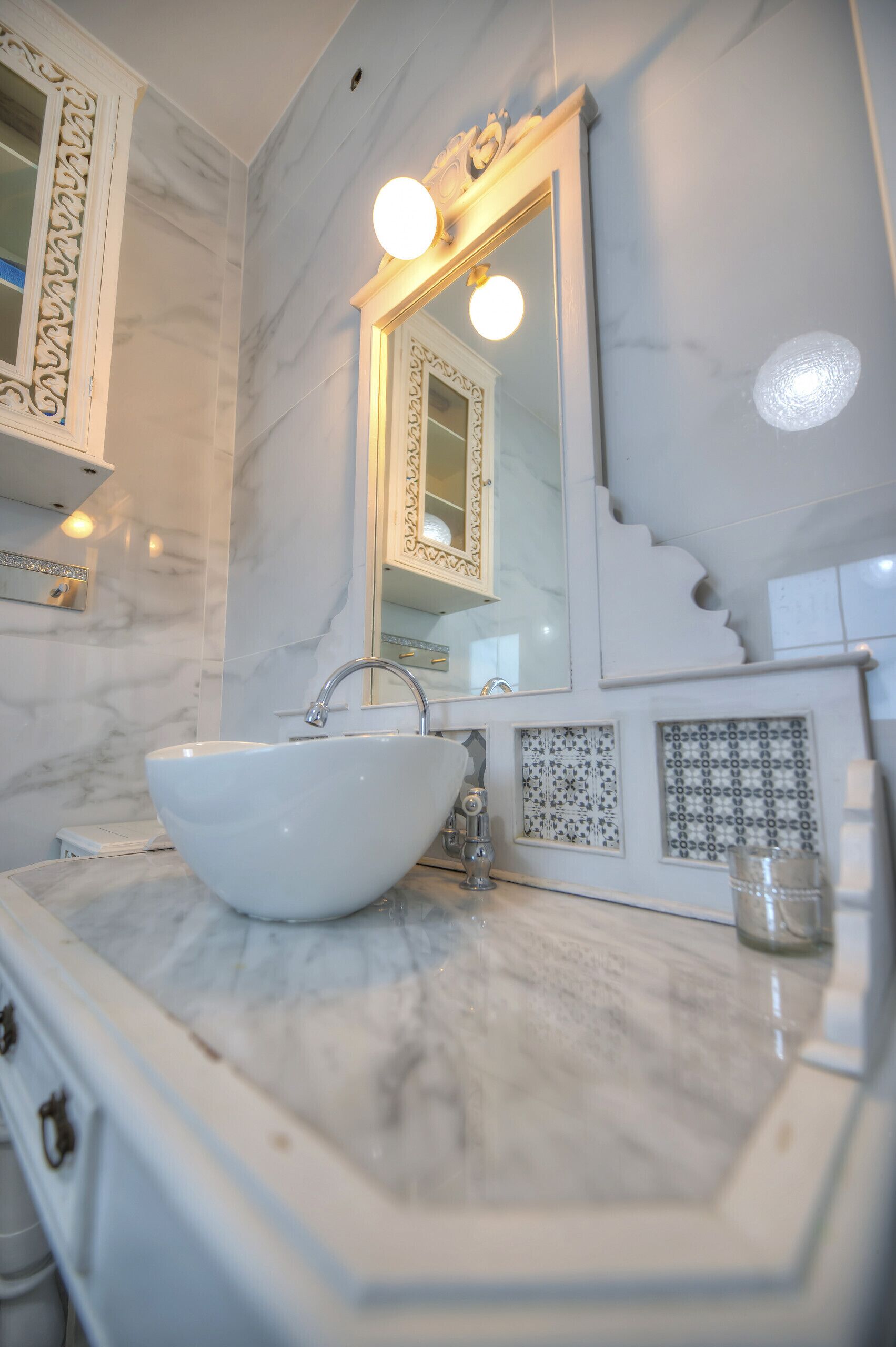 Un baño con lavamanos de mármol blanco, un espejo grande con iluminación y azulejos decorativos.
