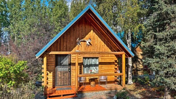 A wooden cabin with a pitched roof, a porch with chairs, and a window with curtains. - Joy's Kenai River Cabins (Sterling)