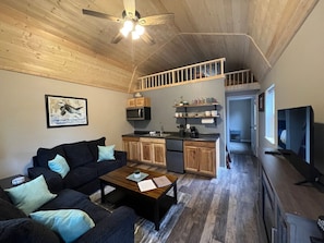 A compact living space with a sofa, coffee table, kitchen area, and a TV. - Mt. Alice Cozy cabin #2 of #3 built new in 2023. (Seward)