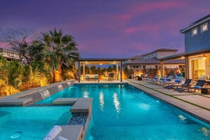Une piscine dotée de chaises longues et d’un espace salon couvert, le tout entouré de palmiers et surplombant un ciel au coucher du soleil.