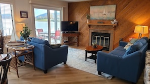 A living room with a fireplace, blue sofas, a TV, and a view of a lake. - The Woodlands, Bernard's Landing, Smith Mountain Lake (Moneta)
