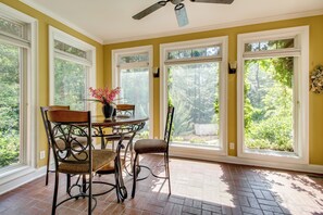 A sunlit room with a dining table, chairs, and large windows offering a view of greenery.