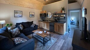 Een gezellige woonkamer met een donkere bank, een salontafel en een keukenhoek met houten kasten en een magnetron.