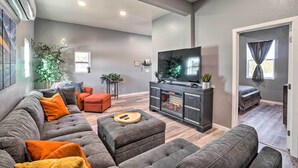Ein modernes Wohnzimmer mit einer grauen Couch, orangefarbenen Kissen, einem Holzfußhocker und einem Fernseher auf einem dunklen Schrank.