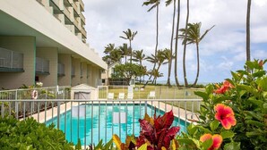 Zona de piscina de hotel con vista despejada al océano, rodeada de palmeras y un edificio con balcones.