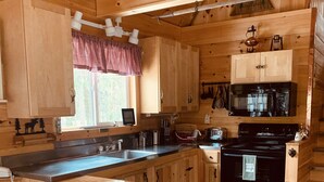 Une cuisine rustique aménagée dans une cabane en bois, avec un îlot central, deux tabourets, un four à micro-ondes, une cuisinière et une fenêtre agrémentée d’un rideau.
