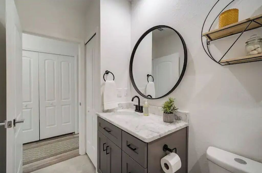 Une salle de bain avec un meuble-lavabo blanc, un miroir rond, des toilettes et une petite étagère contenant des articles de toilette.