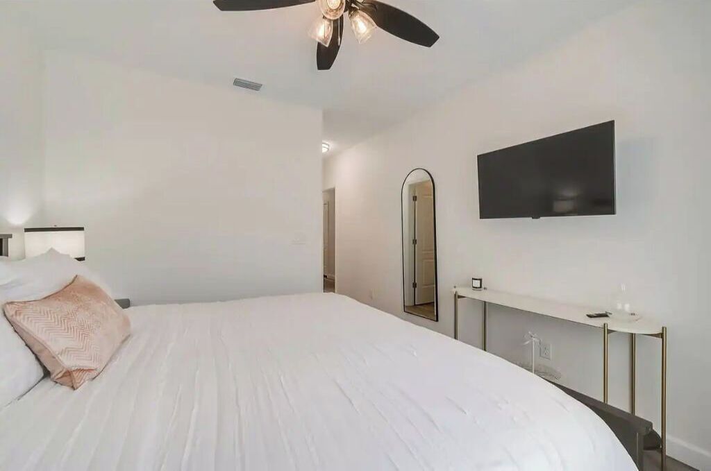 Une chambre avec un grand lit, un ventilateur de plafond, un téléviseur fixé au mur, une table de chevet et un miroir.