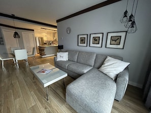 A modern living room with a grey sofa, a glass coffee table, and a dining area with white chairs. - Ski in/ Ski out Modern 1 bedroom with fireplace (Mont-Tremblant)