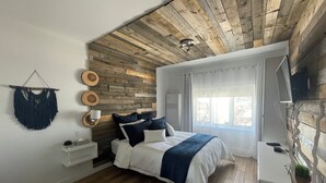 A bedroom with a wooden ceiling, a bed with white bedding, a bedside table, a TV, and a window with curtains. - Ski in/ Ski out Modern 1 bedroom with fireplace (Mont-Tremblant)